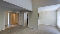 This two-story residence is defined by its impressively vaulted ceilings in the main living area, which create a dramatic sense of vertical space and openness. The living room is carpeted and flows naturally toward adjacent areas, giving the floor plan an open, connected feel. A partial wall divides the living space from the dining area, which is distinguished by an elegant chandelier that adds a refined touch to the otherwise neutral, understated interior. The dining space also features a breakfast bar or pass-through counter, conveniently connecting to the kitchen. The kitchen is visible from the main living area and is equipped with dark appliances, including a refrigerator, along with warm wood-toned cabinetry. A hallway extends toward additional rooms, including what appears to be bedrooms and a bathroom, with multiple doors visible along the corridor. The neutral gray and beige color palette used throughout the walls and carpeting contributes to a cohesive, calm ambiance. White trim and baseboards frame each room cleanly, adding a crisp contrast to the soft tones of the space. The two-story design adds architectural interest and functionality to the overall layout.