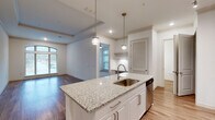 Located at 2925 Buford Dr in Buford, Georgia, this single-floor unit offers a thoughtfully designed open-concept layout with contemporary finishes throughout. The kitchen serves as the centerpiece of the living space, anchored by a substantial granite-topped island with an undermount sink and pendant lighting overhead. White shaker cabinetry reaches toward the ceiling, complemented by a decorative hexagonal tile backsplash and a full suite of stainless steel appliances, including a French door refrigerator, electric range, and over-the-range microwave. A stacked washer and dryer unit is conveniently integrated nearby.

The open living and dining area features hardwood-style flooring and a tray ceiling with crown molding, adding architectural definition to the space. A built-in desk nook provides a dedicated workspace, while an arched window and additional large windows flood the area with natural light and exterior views.

The bedrooms are carpeted for comfort, with one featuring corner windows that maximize daylight and outdoor views. A ceiling fan is present in the bedroom, which connects to a private bathroom. The bathroom includes a white shaker vanity, quartz countertop, a framed mirror, and hardwood-style flooring, maintaining the clean, modern aesthetic found throughout the entire unit.