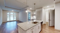Located at 2925 Buford Dr in Buford, Georgia, this single-floor unit offers a thoughtfully designed open-concept layout with contemporary finishes throughout. The kitchen serves as the centerpiece of the living space, anchored by a substantial granite-topped island with an undermount sink and pendant lighting overhead. White shaker cabinetry reaches toward the ceiling, complemented by a decorative hexagonal tile backsplash and a full suite of stainless steel appliances, including a French door refrigerator, electric range, and over-the-range microwave. A stacked washer and dryer unit is conveniently integrated nearby.

The open living and dining area features hardwood-style flooring and a tray ceiling with crown molding, adding architectural definition to the space. A built-in desk nook provides a dedicated workspace, while an arched window and additional large windows flood the area with natural light and exterior views.

The bedrooms are carpeted for comfort, with one featuring corner windows that maximize daylight and outdoor views. A ceiling fan is present in the bedroom, which connects to a private bathroom. The bathroom includes a white shaker vanity, quartz countertop, a framed mirror, and hardwood-style flooring, maintaining the clean, modern aesthetic found throughout the entire unit.