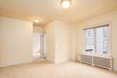 This single-floor space is designed with a straightforward, functional layout spread across multiple interconnected rooms. The living areas are finished with neutral, light beige wall-to-wall carpeting that lends a clean, cohesive look throughout. Walls are painted in a soft warm yellow tone, complemented by white baseboards and trim, contributing to a bright and airy atmosphere. Flush-mount brass-finished ceiling fixtures provide consistent overhead lighting across the carpeted rooms, which connect through white-framed doorways. The layout, visible in the 3D model, reveals a series of rooms arranged in an open, accessible flow.

The kitchen is compact yet practical, featuring white cabinetry extending to the ceiling for generous storage, a gas range, a full-size refrigerator, a single-basin sink, and a window that allows natural light and views of surrounding buildings. The flooring transitions to speckled terrazzo tile in the kitchen and bathroom.

The bathroom includes a vanity with white cabinetry, a countertop sink, a wall-mounted mirror with overhead lighting, a window for natural ventilation, and a separate shower or tub area finished in white tile. One room features a radiator unit beneath a window with urban views, indicating a traditional heating system throughout the space.