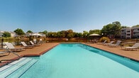 Located in Southaven, Mississippi, this outdoor pool area serves as a well-appointed community amenity designed for relaxation and recreation. The centerpiece is a generously sized rectangular swimming pool with clear, blue-tinted water, framed by brick-accented coping and a wide surrounding deck finished in textured concrete. The expansive sun deck provides ample space for lounging, furnished with multiple chaise lounge chairs arranged along all sides of the pool, as well as several patio umbrella stations offering shaded seating areas for those seeking relief from the Mississippi sun. The pool area is fully enclosed by a black wrought iron fence, ensuring both security and an open, airy feel that allows views of the surrounding mature tree line and neighboring greenery. A charming poolside structure with a pitched shingle roof, large windows, and siding exterior anchors one end of the pool deck, offering an accessible amenity building for residents. The overall atmosphere is bright and inviting, benefiting from abundant natural sunlight and clear open skies. The well-maintained grounds and thoughtful layout make this pool area an ideal outdoor retreat for the community it serves.