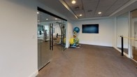 This yoga and spin studio is a thoughtfully designed, single-floor fitness space that seamlessly blends multiple training disciplines into one cohesive environment. The main studio area features durable carpet flooring and clean white walls, creating a bright and motivating atmosphere. A wall-mounted flat-screen TV serves as a central focal point, ideal for guided workouts or virtual classes. Floor-to-ceiling mirrors line one wall, amplifying the sense of space and allowing users to monitor their form during movement.

A freestanding ballet barre runs across the studio, supporting yoga, barre, and stretching routines. Spin bikes are positioned near the windows, which offer views of the surrounding greenery and bring in natural light. A well-stocked medicine ball rack adds functional training versatility to the space.

Elegant glass partition walls with black framing separate the studio from an adjacent fitness area equipped with cardio machines, including treadmills, and strength training equipment. This transparent division maintains an open, airy feel while clearly defining each functional zone. The drop ceiling with recessed lighting provides consistent, even illumination throughout.

Together, the two connected spaces form a comprehensive fitness facility suited for a wide range of activities, from low-impact yoga and barre to high-intensity cycling and strength training.