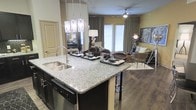 This single-floor residence offers a seamlessly integrated open-concept design that connects the kitchen and living areas into one cohesive, functional space. The kitchen serves as the centerpiece, anchored by a large granite-topped island with a stainless steel sink, dark cabinetry, and a built-in dishwasher. Elegant pendant lighting hangs above the island, adding a refined touch to the space. Hardwood-style flooring extends throughout, lending warmth and continuity to the layout. The living area is generously sized, featuring a ceiling fan and floor-to-ceiling curtains that frame large windows and glass doors, allowing abundant natural light to fill the room. The neutral color palette of warm taupes and soft yellows creates an inviting, contemporary ambiance throughout. Decorative wall art and thoughtfully placed accent lighting contribute to the polished, curated aesthetic. The open sightlines between the kitchen and living space make this residence ideal for both everyday living and entertaining, offering a comfortable and visually cohesive environment from every angle.