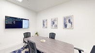 Located at 1025 E 25th St in Hialeah, FL, this single-room office space offers a polished and functional environment designed for productive meetings and collaboration. The room is thoughtfully appointed with crisp white walls that enhance the bright, open atmosphere, complemented by light-toned tile flooring and a bold navy and gold area rug that anchors the central conference table. The elongated gray conference table is surrounded by ergonomic mesh task chairs, comfortably accommodating a small group for focused discussions or presentations. A large wall-mounted flat-screen display serves as a key focal point, ideal for presentations, video conferencing, or digital showcasing. A skylight overhead allows natural light to filter in, adding warmth to the clean, contemporary aesthetic. Framed abstract artwork in coordinating blue and gray tones adorns one wall, contributing to the professional yet welcoming ambiance. Full-height glass partition doors with a white frame open the space to a larger communal area beyond, providing both visual connectivity and the ability to maintain privacy when needed. A small accent stool adds a subtle touch of warmth. This well-designed office space delivers a streamlined, modern setting ideal for professional use in the heart of Hialeah.