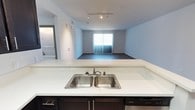 This single-floor unit showcases an open-concept layout that seamlessly connects the kitchen and living area, creating a cohesive and functional living space. The kitchen is equipped with a double-basin stainless steel sink set into a clean white countertop, complemented by dark cabinetry beneath and a dishwasher. The peninsula-style counter design allows for an unobstructed sightline into the living area, making the space feel expansive and connected.

Track lighting runs along the ceiling, providing adjustable illumination throughout the main living zone. The living area features dark flooring that contrasts with the light-toned walls, adding depth and visual interest to the space. A large window with vertical blinds anchors the far wall of the living area, allowing natural light to filter in.

The soft, neutral blue-grey walls throughout contribute to a calm and modern ambiance. A separate area, visible through a doorway on the left, appears to house a stacked washer and dryer, adding practical in-unit laundry convenience. Overall, the space presents a clean, contemporary design with an efficient and open feel.