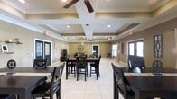 This single-floor gathering space offers a polished and functional environment designed for communal use. The room is defined by its distinctive coffered ceiling, which adds architectural depth and visual interest overhead. Ceiling fans are integrated into the coffered design, providing both style and practical airflow throughout the space. Recessed lighting complements the natural light that filters in through multiple windows and glass-paneled doors, creating a bright and inviting ambiance. The large-format light tile flooring ties the room together and enhances the sense of openness. Multiple dark-stained wood dining sets are arranged throughout the space, accommodating a variety of group sizes and making it ideal for gatherings, meetings, or casual dining. The warm tan and neutral wall tones lend a cozy yet refined feel to the interior. Decorative touches, including a large round wall clock and floral accents, add personality to the space. Several entry points with dark-framed doors provide convenient access from multiple directions. Overall, this space strikes a balance between elegant design and practical functionality, making it a versatile venue suited to a wide range of social activities.