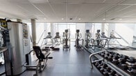 Located in the heart of downtown St. Louis at 411 N Eighth St, this well-appointed fitness center offers a comprehensive workout environment designed for both cardio and strength training. The space is defined by floor-to-ceiling windows that flood the interior with natural light while offering striking views of the St. Louis skyline, creating an energizing atmosphere for exercise. The cardio zone is equipped with multiple treadmills, elliptical trainers, and additional aerobic machines, all arranged to take full advantage of the panoramic city views. Full-length mirrored walls run throughout the space, visually expanding the room and supporting proper form during workouts. The strength training area features a robust selection of dumbbells on a dedicated rack, adjustable weight benches, and a cable/multi-use resistance machine, providing a thorough free-weight and machine training experience. Stability and balance tools, including exercise balls and a BOSU trainer, are also available for functional fitness routines. The flooring transitions between light-toned rubber matting in the cardio zone and darker, impact-absorbing rubber in the free weights area. Recessed ceiling lighting complements the natural light, while the clean white walls and minimalist design maintain a professional and motivating environment throughout.