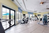 Located in Hialeah, FL, this single-floor fitness and recreation facility offers a generously sized, well-appointed space designed to accommodate a wide range of activities. The main fitness area is bright and airy, bathed in natural light streaming through large multi-pane windows that line the walls and offer views of the surrounding greenery. The ceiling features elegant tray detailing with recessed lighting and multiple ceiling fans that ensure comfortable airflow throughout. The room is finished with durable large-format tile flooring and painted in soft yellow and blue tones, creating an inviting atmosphere. It is equipped with a comprehensive array of cardio machines—including treadmills, ellipticals, and stair climbers—as well as strength training equipment and cable machines. An adjoining cycling studio, styled with calming blue walls, houses a row of stationary bikes, wall-mounted mirrors, and its own ceiling fan, providing an ideal environment for group fitness sessions. A separate children's play area offers a cheerful, imaginatively decorated space with vivid nature-themed murals, colorful striped flooring, a crawl-through play tunnel, and a wall-mounted television, making it a safe and engaging area for younger visitors. Together, these spaces form a comprehensive community wellness facility.