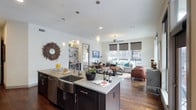 This well-appointed 2-bedroom, 2-bathroom residence offers a thoughtfully designed single-floor layout that maximizes both comfort and functionality. The open-concept living area serves as the heart of the home, seamlessly connecting the kitchen, dining, and living spaces for an expansive, airy feel.

The modern kitchen is anchored by a large center island with a deep farmhouse-style sink, quartz countertops, and dark espresso cabinetry. Stainless steel appliances and pendant lighting above the island add a polished, contemporary touch. The kitchen flows effortlessly into the dining and living areas, making it ideal for entertaining.

The living room is bathed in natural light courtesy of floor-to-ceiling windows dressed with sleek roller shades, offering views of the surrounding exterior. Rich hardwood flooring runs throughout the main living area, complementing the neutral, light-toned walls. A ceiling fan adds both style and practicality to the space.

Built-in shelving in an adjacent nook provides additional storage and display space, adding character and versatility. The overall ambiance is modern and clean, with a warm undertone created by the wood flooring and dark cabinetry accents. This residence combines generous proportions with upscale finishes for a refined everyday living experience.