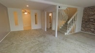This three-floor residential space features an open main living area finished with wall-to-wall beige carpeting, creating a neutral, cohesive aesthetic throughout. The layout is defined by a structural support column that subtly divides the space while maintaining an open feel. A ceiling fan with a light fixture is installed overhead in one section of the living area, providing both air circulation and ambient lighting, complemented by recessed lighting in the ceiling. White-painted walls and trim contribute to a clean, bright interior atmosphere. A staircase with a white wood banister is positioned along one side of the main level, providing access to the upper floors. Toward the rear of the main level, an open doorway leads to what appears to be a utility or laundry area, visible by the appliances within. A secondary room or alcove is also accessible from the main living space, featuring its own lighting. HVAC vents are integrated into the ceiling, indicating a central air system. The overall design is straightforward and functional, offering a blank canvas adaptable to various furnishing arrangements across its multiple levels.