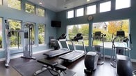 This fitness center offers a bright and inviting environment designed to support a well-rounded workout routine. The room is finished with cool blue-gray walls that contribute to a calm, focused atmosphere, complemented by dark hardwood-style flooring that adds a clean, contemporary look throughout.

The space is generously windowed, with large lower-level windows and a row of clerestory windows above, flooding the room with natural light and providing open views of lush green landscaping and surrounding outdoor scenery. This abundant glazing creates an airy, energizing environment ideal for exercise.

Cardio equipment is prominently featured, including multiple treadmills with individual console displays lined along the windowed wall, an upright elliptical trainer, and an additional upright cycle stationed nearby. A weight bench and cable resistance machine are also present, offering options for strength training alongside the cardio offerings. A wall-mounted flat-screen television provides entertainment during workouts, while a wall clock keeps users on schedule. Recessed ceiling lighting supplements the natural light, ensuring the space remains well-lit throughout the day.