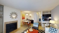 Located at 2134 South Richfield Way in Aurora, Colorado, this single-floor residence offers a thoughtfully designed open-concept layout that seamlessly connects the living, dining, and kitchen areas. The living room serves as a welcoming focal point, anchored by a tiled fireplace and decorated with stylish accents including a sunburst mirror and bold geometric area rug. Plush carpeting runs throughout the main living areas, adding warmth and comfort underfoot. The dining area sits adjacent to the living space and is illuminated by a contemporary pendant light, with seating for several guests and a vibrant orange abstract artwork that adds a pop of color to the neutral walls. A white buffet cabinet provides additional storage and display space. The kitchen is visible from the main living area and features dark cabinetry with modern hardware, a tiled backsplash, and bar-height seating, making it ideal for casual dining and entertaining. A hallway leads to additional rooms, including what appears to be a bedroom and bathroom, maintaining privacy while keeping the layout efficient. The textured ceiling and neutral wall tones provide a clean, versatile backdrop throughout the home, making the space easy to personalize.