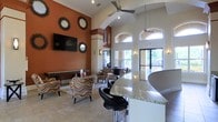 This single-story space makes a strong architectural impression through its dramatic vaulted ceilings, which lend an expansive, airy quality to the interior. A bold terracotta-orange accent wall serves as a focal point, framing a wall-mounted flat-screen television and flanked by decorative sunburst mirrors that add a stylish, contemporary touch. Ceiling fans are positioned throughout to complement the open volume of the space.

The lounge area features a comfortable seating arrangement of multicolored patterned armchairs arranged on a neutral area rug, creating an inviting gathering zone. A granite-topped bar counter with black leather bar stools defines a separate service or reception area, adding functionality and a polished finish to the space.

Large arched windows along the far wall flood the interior with natural light while offering views of an outdoor area beyond, enhancing the sense of openness. Tile flooring in a warm neutral tone runs throughout, tying the design elements together cohesively. Wall sconces provide ambient accent lighting, contributing to an overall atmosphere that is both welcoming and well-appointed.