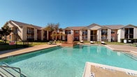 This community pool area offers a welcoming and well-designed outdoor retreat. The centerpiece is a generously sized swimming pool with clear, blue water and a striking brick water feature at its center, creating a resort-like atmosphere. The pool deck provides ample space for lounge chairs and outdoor seating, ideal for relaxation under open skies. The area is enclosed by a black iron security fence, ensuring both safety and a clean aesthetic.

Adjacent to the pool is a covered pavilion structure featuring elegant white columns framing a graceful arched entryway, offering shaded views directly overlooking the pool. This covered area serves as an inviting gathering space for residents.

A separate pool house or clubhouse structure is accessible via full-length French-style glass doors, which allow abundant natural light to filter through while maintaining a visual connection to the pool area. The interior of this building features an octagonal concrete floor plan with multiple entry and window points, lending the space a unique architectural character. Additional covered porch areas with large windows and horizontal lap siding provide supplementary outdoor lounge space. The surrounding grounds include mature trees and well-maintained lawns, completing the serene, community-focused setting.