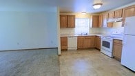 This single-story space is designed with practicality and comfort in mind. The kitchen is a standout feature, outfitted with warm medium-toned wood cabinetry offering generous upper and lower storage. Light-colored countertops provide ample workspace, and a window above the sink allows natural light to brighten the area. White appliances, including a dishwasher, range, and microwave, are neatly integrated into the layout. The kitchen floor is finished with neutral tile in a classic pattern, adding a clean, easy-to-maintain surface underfoot. A structural column defines the boundary between the kitchen and the adjoining living area, which transitions to wall-to-wall carpet, creating a softer, more comfortable zone for relaxation. Flush-mount ceiling light fixtures provide consistent overhead lighting throughout. The overall color palette is neutral and understated, with soft wall tones complementing the wood cabinetry and flooring. The open sightlines between the kitchen and living area contribute to a connected, functional layout well-suited to everyday living.