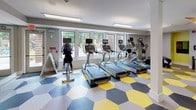 This fitness center is a thoughtfully designed workout facility that combines functional layout with energetic aesthetics. The space is immediately defined by its striking hexagonal-patterned floor in yellow, gray, and charcoal tones, adding a bold visual identity throughout the room. The facility is generously illuminated by a row of large windows that invite natural light and offer views of surrounding greenery, creating an open and motivating atmosphere. Overhead, recessed lighting and hanging linear fixtures ensure consistent brightness across the entire space, complemented by a ceiling fan that promotes airflow. The cardio section is well-stocked with multiple treadmills, elliptical trainers, and stationary bikes, each equipped with personal digital screens. A dedicated free weights area features a tiered dumbbell rack alongside an adjustable weight bench, ideal for strength training. Full-length mirrors line one wall, enhancing the sense of depth and allowing users to monitor their form during workouts. Accent walls in a vivid chartreuse green provide pops of color against the neutral gray walls. A staircase access point is visible within the space, and structural columns are integrated cleanly into the layout. The overall ambiance is modern, energetic, and purpose-built for an active lifestyle.