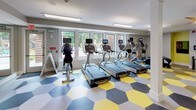 This fitness center is a thoughtfully designed workout facility that combines functional layout with energetic aesthetics. The space is immediately defined by its striking hexagonal-patterned floor in yellow, gray, and charcoal tones, adding a bold visual identity throughout the room. The facility is generously illuminated by a row of large windows that invite natural light and offer views of surrounding greenery, creating an open and motivating atmosphere. Overhead, recessed lighting and hanging linear fixtures ensure consistent brightness across the entire space, complemented by a ceiling fan that promotes airflow. The cardio section is well-stocked with multiple treadmills, elliptical trainers, and stationary bikes, each equipped with personal digital screens. A dedicated free weights area features a tiered dumbbell rack alongside an adjustable weight bench, ideal for strength training. Full-length mirrors line one wall, enhancing the sense of depth and allowing users to monitor their form during workouts. Accent walls in a vivid chartreuse green provide pops of color against the neutral gray walls. A staircase access point is visible within the space, and structural columns are integrated cleanly into the layout. The overall ambiance is modern, energetic, and purpose-built for an active lifestyle.