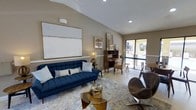 This single-story clubhouse is thoughtfully designed to provide a comfortable and stylish communal gathering space. The main lounge area features vaulted ceilings with recessed lighting, contributing to an open and airy atmosphere. Warm neutral wall tones are complemented by light-colored flooring, creating a cohesive and inviting aesthetic throughout.

The lounge is furnished with a bold navy blue tufted sofa accented by patterned throw pillows, anchored by a colorful area rug and a glass-top coffee table. A sculptural accent chair in rich brown leather adds a contemporary touch, while a vintage-style wooden secretary desk and decorative cabinet provide functional charm. A large abstract canvas artwork adorns one wall, serving as a striking focal point.

Natural light flows in through a sliding glass door that offers views of the exterior surroundings, brightening the space and creating a seamless connection to the outdoors. A bistro-style seating area near the window provides an intimate spot for conversation or casual work. Decorative accessories, including a table lamp and curated art pieces, add warmth and personality. Overall, the clubhouse combines modern design elements with comfortable furnishings to create a versatile and welcoming environment for residents.