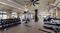 Located at 8205 Commerce Way in Miami Lakes, Florida, this fitness center offers a generously sized, single-floor layout designed to support a full range of workout routines. The main exercise room is defined by its open, airy feel, supported by large floor-to-ceiling windows that flood the space with natural light and provide views of the surrounding outdoor greenery. Textured stone accent walls and bold dark structural columns add a modern, upscale aesthetic throughout.

The exercise room is outfitted with an extensive selection of equipment, including a full dumbbell rack, cable machines, weight benches, treadmills, stair climbers, and stationary bikes, catering to both strength training and cardiovascular fitness. Oversized wall mirrors amplify the sense of space and allow for proper form monitoring. Durable rubber flooring covers the workout areas, while large ceiling fans and recessed lighting ensure comfort during exercise.

A separate, glass-walled studio room with frosted panels offers a quieter space ideal for yoga, stretching, or personal training sessions. The facility also features a well-appointed entry area with built-in cabinetry and wall-mounted displays. The combination of refined design elements and comprehensive equipment makes this fitness center a standout amenity.