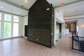 This dedicated yoga room offers a thoughtfully designed environment that balances natural beauty with purposeful simplicity. The space is defined by its soaring ceilings, which lend an expansive, open quality that encourages ease of movement and a sense of calm. Large floor-to-ceiling windows, including a signature arched window, wrap around multiple walls and flood the interior with abundant natural light while framing sweeping views of surrounding woodland. The lush greenery visible through the glass creates an immersive connection to nature, enhancing the meditative atmosphere of the room. Smooth, warm-toned hardwood flooring runs throughout, providing a clean and resilient surface ideal for practice. A dramatic full-height reclaimed wood accent wall serves as a striking focal point, introducing rich texture and an earthy, rustic contrast to the otherwise crisp, neutral walls and trim. Recessed ceiling lighting supplements the generous natural light, ensuring the space remains well-lit at any time of day. The open, uncluttered layout provides ample room for individual or group practice, and thoughtful details such as wall-mounted mat storage demonstrate the space's dedicated functionality. Overall, the room exudes a tranquil, grounded ambiance perfectly suited to yoga, meditation, and mindful movement.