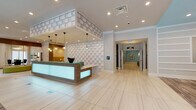 Located on E Madison Street in Tampa, Florida, this single-floor leasing and amenity space presents a contemporary design aesthetic defined by light wood-look tile flooring, teal and aqua accent tones, and thoughtfully curated artwork throughout. The entrance welcomes visitors through floor-to-ceiling glass doors that flood the space with natural light, revealing a striking striped architectural column and a sophisticated seating lounge with a starburst chandelier, plush navy chairs, and views to the surrounding landscape. The reception area anchors the space with an illuminated backlit desk, textured 3D wall panels, and pendant globe lighting that create a polished first impression. A dedicated conference room features large windows overlooking the streetscape, a statement chandelier, teal ceiling detail, and a full boardroom table with contemporary seating. The co-working lounge offers dual workstations with mounted displays, built-in shelving, and elegant crystal-style pendant fixtures beneath a teal tray ceiling. Additional lounge areas provide casual seating with vibrant upholstered benches and sculptural art installations. The elevator lobby is finished with tray ceilings, soft blue walls, framed artwork, and a tufted bench, rounding out a cohesive, design-forward environment.