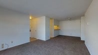 This single-floor residence offers a straightforward, functional layout designed for comfortable everyday living. The main living area is finished with wall-to-wall carpet in a neutral tone, providing a warm and quiet underfoot experience. A textured popcorn ceiling runs throughout, adding subtle visual depth to the space. The living area flows openly, with a ceiling fan providing air circulation and a practical touch to the room's design. A partial dividing wall separates the living space from the kitchen area, which features a pass-through counter — an ideal setup for casual dining or easy serving between rooms. The kitchen is warmly lit, with under-cabinet lighting visible above the counter, creating an inviting cooking environment. Tile flooring in the adjacent entry and hallway areas provides a durable transition zone from the main living space. A hallway leads to additional rooms, with a white door visible at the end, suggesting a private area beyond. The unit's neutral white walls throughout offer a clean, versatile backdrop well-suited to a variety of décor styles. The overall ambiance is calm and understated, offering a practical and livable single-floor layout.