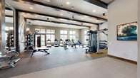 Located at 1400 N Main St in Keller, TX, this fitness center offers a generously sized, single-floor exercise space designed for a comprehensive workout experience. The open layout accommodates a wide variety of equipment, including treadmills, rowing machines, cycling bikes, a StairMaster, cable machines, and a full dumbbell rack — providing options for both cardio training and strength conditioning.

Architecturally, the space features distinctive dark exposed beams set against a light, neutral ceiling, lending a modern yet grounded aesthetic. Recessed lighting and ceiling fans are distributed throughout, ensuring both visibility and comfort during workouts. Large windows along the perimeter walls flood the interior with natural light and offer outdoor views, creating an open and energizing atmosphere.

A standout design element is the accent wall featuring large framed mirrors paired with geometric, backlit lighting in a criss-cross pattern — enhancing the sense of space while adding a contemporary visual flair. The flooring transitions between durable carpet tile and smooth hard surface zones, defining distinct workout areas. A built-in wooden shelving unit with a wall-mounted TV and counter space provides a functional amenity station. The high ceilings and expansive floor plan collectively create a professional, well-equipped fitness environment suited for a full range of exercise routines.