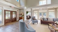 This single-floor space is designed with an open, flowing layout that seamlessly connects multiple functional areas. The grand entrance foyer immediately impresses with soaring two-story ceilings, transom windows that flood the space with natural light, and large French doors framing views of the surrounding landscape. Decorative columns define transitions between zones without interrupting the open feel.

The central living area features large-format tile flooring that extends throughout the main corridors, transitioning to plush carpet in the seating zones. A standout stone-surround fireplace anchors the room, topped by a rich dark wood mantel and built-in cabinetry housing a wall-mounted television. Multiple seating arrangements accommodate both intimate conversation and larger gatherings.

A secondary lounge or office area is accessible via a connecting hallway, offering a quieter retreat with carpeted flooring, framed wall art, and ample natural light from side windows. Additional areas are accessible along a well-lit corridor leading to private rooms.

Throughout the space, neutral gray and warm beige tones create a cohesive, welcoming ambiance. Recessed lighting, crown molding, and carefully placed floor lamps contribute to a polished, comfortable atmosphere suited for a variety of uses.