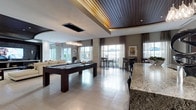 This single-floor amenity space is designed with a contemporary aesthetic, combining functionality and upscale finishes throughout. The open-plan layout accommodates multiple distinct zones, creating a versatile environment suited for both socializing and recreation. Large windows dressed with floor-length drapes flood the interior with natural light, offering views of the surrounding exterior.

A centerpiece dark-finished pool table anchors the recreational zone, positioned beneath a striking pendant chandelier that adds a refined focal point. The ceiling design is a notable architectural feature, incorporating warm wood slat paneling inset into a recessed tray detail, creating visual depth and contrast against the white surrounding surfaces.

Large-format light-toned porcelain tiles span the floor, contributing to the bright, airy atmosphere. A granite-topped bar counter occupies the foreground, providing a practical surface for entertaining. Adjacent high-top tables with bar stools offer casual dining and gathering space near the windows.

A comfortable lounge seating area with neutral upholstered sofas is visible in the background, completing the multi-functional layout. The overall ambiance is polished and inviting, balancing modern design elements with warm material accents.