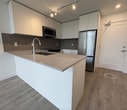 Located at 14575 104 Ave, Unit 620 in Surrey, BC, this 450-square-foot, one-bedroom, one-bathroom condo delivers a modern and efficient living experience across a single floor. The open-concept kitchen and living area forms the heart of the unit, featuring wood-look laminate flooring throughout, crisp white walls, and large floor-to-ceiling windows that flood the space with natural light and offer views of the surrounding greenery. The kitchen is outfitted with sleek white cabinetry, a stone countertop peninsula, a tile mosaic backsplash, stainless steel appliances including an over-the-range microwave, and track lighting overhead. The living room benefits from impressively soaring ceilings that amplify the sense of space, along with baseboard heating and expansive windows. The bedroom is a bright, cozy retreat with its own large window framing outdoor views and a tray ceiling detail adding architectural interest. The bathroom features clean, minimalist design with full-height tile surround, a soaker tub, and a toilet. Clean lines, neutral tones, and quality finishes define the overall aesthetic of this well-appointed urban condo.