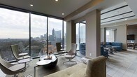 This refined single-floor residence offers a seamless blend of modern design and urban living at its finest. The living area is anchored by expansive floor-to-ceiling glass windows that frame breathtaking panoramic views of a vibrant city skyline, flooding the interior with abundant natural light throughout the day. The neutral, warm-toned walls and ceilings complement the contemporary aesthetic, creating a sophisticated yet inviting atmosphere. Sleek recessed lighting adds a polished finish to the ceiling, enhancing the space after dark. The open layout of the living area is thoughtfully arranged, featuring a plush sofa, a sculptural low-profile coffee table, and a pair of modern accent chairs positioned to take full advantage of the stunning skyline vistas. Large structural columns are elegantly integrated into the design, adding architectural character without interrupting the flow of the space. A glimpse into the adjoining area reveals a continuation of the contemporary palette, with additional seating and decor that maintain a cohesive design language throughout. The combination of high-end finishes, generous proportions, and remarkable city views makes this space a standout urban retreat.