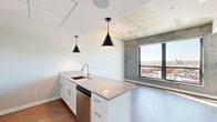Located at 127 N 17th St in Richmond, VA, this one-bedroom, one-bathroom unit offers a thoughtfully designed open-concept layout that maximizes its efficient footprint. The combined kitchen and living area serves as the heart of the space, anchored by a granite-topped island with an undermount sink and pendant lighting. The kitchen features white shaker cabinetry, a white subway tile backsplash, stainless steel appliances including a range with wall-mounted hood, dishwasher, and refrigerator, all set atop durable wood-look flooring. Industrial character is introduced through exposed concrete ceilings and accent walls, lending an urban loft aesthetic throughout. Large windows and a glass balcony door flood the living area with natural light while offering elevated panoramic views of the Richmond cityscape. The bedroom is a comfortable, well-lit retreat with a spacious closet, exposed concrete ceiling detail, and a large window framing the same impressive city views. In-unit laundry adds everyday convenience. The bathroom includes a full soaking tub and shower combination with clean, modern fixtures. A natural wood entry door with a smart lock completes this contemporary urban residence.