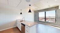 Located at 127 N 17th St in Richmond, VA, this one-bedroom, one-bathroom unit offers a thoughtfully designed open-concept layout that maximizes its efficient footprint. The combined kitchen and living area serves as the heart of the space, anchored by a granite-topped island with an undermount sink and pendant lighting. The kitchen features white shaker cabinetry, a white subway tile backsplash, stainless steel appliances including a range with wall-mounted hood, dishwasher, and refrigerator, all set atop durable wood-look flooring. Industrial character is introduced through exposed concrete ceilings and accent walls, lending an urban loft aesthetic throughout. Large windows and a glass balcony door flood the living area with natural light while offering elevated panoramic views of the Richmond cityscape. The bedroom is a comfortable, well-lit retreat with a spacious closet, exposed concrete ceiling detail, and a large window framing the same impressive city views. In-unit laundry adds everyday convenience. The bathroom includes a full soaking tub and shower combination with clean, modern fixtures. A natural wood entry door with a smart lock completes this contemporary urban residence.