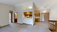 This single-floor residence offers a thoughtfully designed open-concept layout that seamlessly connects the kitchen and living areas. The kitchen is centrally positioned and features warm honey-toned wood cabinetry on multiple walls, providing generous storage space. A peninsula counter creates a natural divide between the kitchen and the carpeted living area, making it ideal for both cooking and entertaining. Vaulted ceilings rise above the space, adding a sense of openness and airiness throughout. A ceiling fan in the living area contributes to the comfortable, functional ambiance. Adjacent to the kitchen, a spacious walk-in closet with built-in shelving offers excellent organizational capacity. A hallway leads to a bathroom, visible in the background, completing the interior layout. The unit also features an outdoor deck with a green-painted wood railing, overlooking a peaceful setting of mature trees and a paved pathway below, providing a private and natural outdoor retreat. The overall aesthetic is clean, neutral, and practical, with light-colored carpeting and white walls that create a bright, welcoming atmosphere throughout the space.