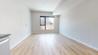 This contemporary single-floor unit showcases a clean, modern design with light-toned hardwood flooring that flows consistently throughout the space, creating a bright and cohesive aesthetic. The open layout connects the living area to the kitchen, maximizing functionality while maintaining an airy feel.

The kitchen is well-appointed with dark cabinetry that provides a striking contrast against the light surroundings, stainless steel appliances including a gas range, dishwasher, and refrigerator, and a stylish marble-patterned tile backsplash. Light stone countertops and a built-in sink complete the functional workspace.

The bedroom area benefits from a large picture window fitted with a roller shade, offering direct views of the surrounding urban skyline, including notable city architecture. Recessed lighting and smooth neutral-toned walls contribute to the room's clean, well-lit atmosphere.

The bathroom features large-format travertine-style floor tiles, a full tub-shower combination with brushed nickel fixtures, a dark-cabinetry vanity with an undermount sink and gray countertop, and a frameless mirror. The neutral palette maintains the unit's consistent modern character throughout. Overall, the space presents a polished, move-in-ready environment suited for contemporary urban living.