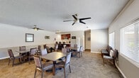 Located at 10453 Artesia Boulevard in Bellflower, California, this single-story clubhouse offers a well-appointed community gathering space designed for comfort and functionality. The main area serves as a versatile dining and social hall, featuring multiple dining tables and upholstered chairs arranged throughout a generously sized, open room. Patterned carpet flooring, textured ceilings, and multiple ceiling fans contribute to a casual and welcoming atmosphere. Wall-mounted air conditioning units and large windows with blinds allow for natural light and climate control throughout the space. A lounge area at one end of the room provides comfortable seating, a wall-mounted flat-screen TV, and a media console, making it suitable for relaxed gatherings or entertainment. The kitchen is efficiently designed with rich dark-wood cabinetry, light granite countertops, a double-basin sink, stainless steel appliances including a microwave and refrigerator with ice and water dispenser, and recessed lighting overhead. A wide kitchen counter doubles as a serving or bar area. A hallway connects to two bathrooms with wood-look flooring and clean, functional finishes. The overall design balances practicality with a comfortable community aesthetic, well-suited for residential gatherings and events.
