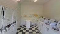 This single-floor shared laundry facility is designed with practicality and efficiency in mind. The room is finished with bright white walls and illuminated by overhead fluorescent lighting, creating a clean and well-lit environment. A bold black-and-white checkered tile floor adds a classic touch to the otherwise utilitarian space.

Along the right wall, a row of top-loading washing machines and dryers are neatly arranged, providing ample capacity for multiple users. On the left side, stacked washer-dryer units make efficient use of vertical space. A centrally positioned white folding table offers a convenient surface for sorting and folding laundry, accompanied by a simple chair for comfort. An informational shelf runs along the back wall, displaying posted notices and instructions to assist users.

The layout is open and easy to navigate, with enough floor space to move comfortably between machines. The overall ambiance is functional and tidy, reflecting a well-maintained communal space suited for residential or multi-unit use.