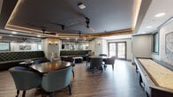 Located in Germantown, Tennessee, this elegantly appointed club room offers a refined social environment designed for both relaxed gatherings and lively entertainment. The space is anchored by warm hardwood-style flooring and framed by tray ceilings with integrated LED accent lighting and wood trim details, lending the room a sophisticated, upscale ambiance. Ceiling fans are strategically placed throughout to enhance comfort.

The lounge area features a richly tufted dark green velvet sofa paired with a distinctive organic-form stone coffee table and a classic upholstered armchair, creating an inviting nook for casual conversation. A large mirror behind the seating visually expands the space.

Multiple seating arrangements include dark wood dining and game tables surrounded by tufted, nailhead-trim chairs in teal and charcoal, offering flexible options for cards, board games, or social dining. A shuffleboard table adds a recreational element along one wall.

The wet bar area is outfitted with sleek cabinetry, marble-look countertops, a mosaic tile backsplash, an under-counter beverage refrigerator, and a large wall-mounted flat-screen TV above, making it an ideal hub for entertaining.

Textured wall coverings and stone accent panels add depth and character throughout, while French doors open to a patio with views of lush greenery, seamlessly connecting the indoors with the outdoors.