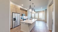 Located at 2080 FM 1102 in New Braunfels, TX, this thoughtfully designed one-bedroom residence offers a clean, modern aesthetic with warm wood-look flooring flowing throughout every room. The open-concept kitchen and living area serves as the heart of the home, anchored by a striking oversized island with a quartz waterfall countertop, an undermount sink, and stylish pendant lighting. Light wood cabinetry extends to the ceiling, maximizing storage, while stainless steel appliances — including a French door refrigerator, range, and microwave — complete the contemporary kitchen. A dedicated laundry closet is conveniently tucked nearby. The living area benefits from abundant natural light through multiple windows dressed with clean white blinds, and a glass-panel door leads directly to the balcony, offering outdoor views of the surrounding landscape. The spacious primary bedroom is well-appointed with dual windows that flood the room with natural light, a ceiling fan, and access to a closet. The neutral, soft-toned walls and recessed lighting throughout create a bright, cohesive ambiance that feels both inviting and functional — ideal for comfortable everyday living in the Texas Hill Country.