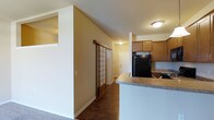 This single-floor residence offers a practical and well-organized layout with clearly defined living areas. The kitchen is positioned toward one side of the space and is equipped with warm honey-toned wood cabinetry, a black refrigerator, and a black range, complemented by light-toned countertops and a white tile backsplash. A peninsula counter extends outward, providing additional workspace and creating a natural boundary between the kitchen and the adjoining area.

One of the more distinctive architectural features of this space is a sliding shoji-style screen partition, which adds a subtle decorative element while offering a flexible room division option. The warm beige walls and flush-mount ceiling lighting contribute to a consistent, neutral aesthetic throughout. Dark hardwood-style flooring runs through the main living and kitchen areas, transitioning to carpet in adjacent spaces, adding textural contrast.

The vaulted ceiling adds a sense of height and openness to the interior, preventing the space from feeling confined. A white panel door at the end of the hallway leads to additional rooms. Overall, the space presents a clean, functional environment with a blend of practical features and understated design details.
