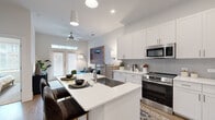 This well-designed one-bedroom, one-bathroom unit offers an efficient and thoughtfully organized single-level layout. The open-concept kitchen and living area form the heart of the space, creating a seamless flow ideal for both daily living and entertaining. The kitchen is appointed with crisp white shaker cabinetry extending to the ceiling, a gray tile backsplash, stainless steel appliances including a French-door refrigerator, electric range, and over-the-range microwave. A central island with an undermount sink and white quartz countertops provides additional prep space and casual seating. Wood-look flooring runs throughout the main living areas, adding warmth to the contemporary design. The living room benefits from abundant natural light through French doors fitted with blinds, and features a ceiling fan overhead. The bedroom is a comfortable retreat, carpeted for comfort, and connects directly to the bathroom. The bathroom is finished with white cabinetry, a quartz vanity top, framed mirror, and a decorative shower curtain, with additional built-in shelving for storage. A dedicated closet completes the unit's practical storage offerings. The overall aesthetic is clean, bright, and modern throughout.