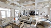 This single-story space showcases a refined, open-concept design highlighted by striking coffered ceilings with white beams set against a warm taupe backdrop, adding architectural depth and grandeur. Recessed lighting throughout, complemented by an elegant chandelier, ensures the space is well-illuminated while maintaining a sophisticated ambiance.

The expansive living area is anchored by a plush neutral-toned sofa arrangement atop a floral area rug, creating a comfortable yet stylish gathering space. Large French doors along one wall flood the interior with natural light while providing an inviting connection to the exterior.

Transitioning seamlessly from the living area, the open layout flows into a kitchen featuring dark cabinetry and stainless steel appliances, offering a bold contrast to the lighter tones throughout the rest of the space. A raised bar with seating bridges the kitchen and living zones, ideal for casual dining or entertaining.

Light hardwood flooring runs consistently throughout, unifying the open layout and enhancing the overall sense of spaciousness. The neutral color palette of whites, grays, and warm beiges creates a cohesive, welcoming atmosphere throughout this thoughtfully designed space.