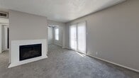 This single-floor, 2-bedroom, 1-bathroom home offers a functional and comfortable layout. The combined kitchen and living room serves as the heart of the space, where a white-painted brick fireplace anchors the living area alongside a sliding glass door that floods the room with natural light. The kitchen is outfitted with white cabinetry, a peninsula with a breakfast bar, light countertops, black appliances including a refrigerator and dishwasher, and wood-look hard flooring that contrasts with the plush carpeting found throughout the rest of the home. Both bedrooms feature neutral gray walls, textured ceilings, and windows with blinds that allow for natural light. The spacious primary bedroom includes a ceiling fan, while the second bedroom also benefits from its own ceiling fan. A dedicated closet with built-in shelving and hanging rods provides generous storage. The bathroom is finished in a modern style, featuring a dual-sink vanity with white quartz countertops, matte black fixtures, round and rectangular mirrors, and wood-look flooring that ties in with the kitchen. A connecting hallway links all rooms together, creating a cohesive and practical flow throughout the home.