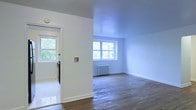 This single-floor space presents a clean, minimalist aesthetic defined by crisp white walls and smooth ceilings that create a bright, open atmosphere. The main living area is finished with rich hardwood flooring that adds warmth and character to the interior. Natural light filters generously through multiple windows, including a wide multi-pane window in the main room that frames views of lush greenery outside, contributing to an airy, welcoming ambiance. A classic cast-iron radiator beneath the window is a notable architectural detail, nodding to timeless building craftsmanship. The entryway, accessible through a solid door with a window panel, features light-toned flooring that transitions seamlessly into the hardwood of the main living space. Clean white baseboards frame each room, providing a polished finishing touch throughout. The layout flows simply and efficiently, with a wall-mounted control panel near the entry adding a practical element. The overall design is understated and versatile, offering a neutral canvas well-suited for a variety of uses and furnishing styles.