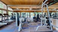 Located at 2600 Bark Wood Rd in Schaumburg, Illinois, this well-equipped fitness center offers a comprehensive workout environment within a single-floor facility. The space is defined by its striking architectural design, featuring soaring vaulted ceilings clad in warm wood paneling, supported by exposed beams and complemented by ceiling fans that maintain a comfortable atmosphere throughout.

Natural light floods the interior through expansive floor-to-ceiling glass windows and sliding doors that line multiple walls, offering refreshing views of the surrounding mature trees and greenery. The open, airy layout is further enhanced by large wall mirrors that amplify the sense of space.

The fitness floor is outfitted with a robust selection of Matrix-branded cardio machines, including multiple treadmills, ellipticals, and a rowing machine, arranged for optimal flow and visibility. Strength training is well represented with a variety of cable machines, weight benches, a squat rack, a full dumbbell rack, and an assortment of medicine balls and stability balls for functional training.

Patterned blue and gray carpet tiles cover the floor, adding both comfort and visual interest. Wall-mounted flat-screen televisions are positioned for easy viewing during cardio workouts. The combination of natural light, lush outdoor scenery, quality equipment, and warm architectural details makes this fitness center an inviting and functional space for a wide range of workout routines.