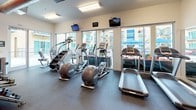 This fitness center offers a comprehensive, single-floor workout facility designed to accommodate a wide range of exercise routines. The space is characterized by a clean, modern aesthetic, featuring a neutral color palette accented by bold orange walls that add energy and visual interest to the environment. Large floor-to-ceiling windows and glass doors flood the interior with natural light and provide open views of the surrounding outdoor area, creating an inviting and motivating atmosphere.

The cardio zone is well-stocked with treadmills, elliptical trainers, and stair climbers, all positioned to take advantage of the outdoor views. Overhead, wall-mounted televisions are conveniently placed throughout the space to keep members engaged during workouts. The strength training area includes a cable machine system and adjustable benches, while a dedicated free weight section offers a full rack of dumbbells for versatile training options. Stability balls, resistance accessories, and additional fitness props are neatly organized on a dedicated rack, adding further variety to available workouts.

Durable dark rubber flooring covers the entire workout area, providing both safety and functionality. Exposed ductwork along the upper walls adds an industrial design element, complementing the open, high-ceiling feel of the space. A utility area near the exit includes storage shelving and a small refrigeration unit, ensuring convenience for users. Direct exterior access through a glass door connects the fitness center to an adjacent outdoor space.