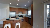 This thoughtfully designed two-floor space combines functionality and modern style throughout. The kitchen serves as a standout feature, showcasing rich warm-toned wood cabinetry that extends from floor to ceiling, offering generous storage. Stainless steel appliances, including a refrigerator, gas range with overhead ventilation hood, and dishwasher, lend a sleek, contemporary feel. Granite-style countertops provide durable workspace, while a central island with an extended counter overhang accommodates casual bar seating, making it ideal for both meal preparation and informal dining. A window above the sink allows natural light to brighten the space, complemented by recessed ceiling lighting that ensures a well-lit environment throughout the day and evening. The kitchen flows seamlessly into the adjacent living areas, supported by warm-toned hardwood flooring that adds continuity and a sense of warmth across the main level. Clean wall lines, neutral grey tones, and modern hardware contribute to the overall contemporary aesthetic. The second floor adds additional living or sleeping accommodations, rounding out this versatile and practical multi-level space.