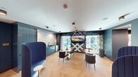 Located at 195 Steely in Irvine, California, this single-floor amenity lounge is a thoughtfully designed communal space that blends contemporary style with functional comfort. The interior is defined by rich teal-painted walls and cabinetry that create a bold, cohesive aesthetic throughout. Underfoot, light-toned herringbone hardwood flooring adds a refined, geometric texture that complements the overall design language.

The main lounge area features a mix of seating arrangements, including round wooden dining tables with modern upholstered chairs finished in warm metallic gold-toned bases, providing casual gathering spots for small groups. A standout design element is the dramatic accent wall, adorned with a graphic black-and-white crosshatch pattern, against which a large wall-mounted flat-screen television is centered, serving as both entertainment and focal point.

Tall, privacy-oriented high-back booth seating upholstered in deep cobalt blue adds a semi-private lounge option ideal for focused work or quiet conversation. A built-in kitchenette along one wall, featuring teal cabinetry, a sink, and under-counter appliances, offers convenient refreshment access. Floor-to-ceiling windows and glass doors flood the space with natural light and provide views of an outdoor area beyond. Lush potted indoor plants and curated wall art contribute warmth and personality, rounding out this inviting and versatile communal lounge.