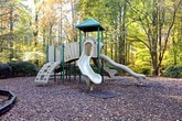 This outdoor playground area offers a welcoming recreational space surrounded by mature deciduous and evergreen trees that provide natural shade and a tranquil, park-like atmosphere. The play area is defined by a bordered perimeter filled with wood chip surfacing, which cushions the ground beneath the central play structure. The multi-level play unit serves as the focal point of the space, featuring multiple slides — including a wide double slide and a single curved slide — along with climbing nets and elevated platforms supported by green metal poles, encouraging active and imaginative play. Scattered throughout the area are green painted benches, thoughtfully positioned to offer comfortable seating for supervising adults. Well-maintained hedges and ornamental shrubs line portions of the perimeter, adding greenery and structure to the landscape. The playground sits within a broader residential setting, with multi-story buildings visible in the background, their balconies and facade contributing to a community-oriented environment. The open layout and abundance of natural surroundings make this a bright and inviting space during daylight hours, with the woodland backdrop enhancing its sense of seclusion and connection to nature.