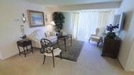 This single-floor residence offers a refined and comfortable living environment defined by classic design sensibilities and a warm, neutral color palette. The living area serves as the centerpiece of the space, showcasing plush, cream-toned upholstered seating including a loveseat and a wingback chair, arranged thoughtfully around a dark wood coffee table atop an ornate area rug. A decorative lattice-back accent chair adds an elegant touch to the arrangement. Large floor-to-ceiling windows fitted with vertical blinds allow abundant natural light to fill the room, creating a bright and airy atmosphere. Wall-to-wall carpeting in soft beige tones contributes to the space's understated warmth. A framed coastal landscape painting adorns one wall, complemented by a table lamp and a lush indoor plant that bring life and character to the interior. A dark wood dresser or console is partially visible, suggesting thoughtful use of storage and furnishing throughout. The overall ambiance is one of quiet elegance — a traditionally styled, light-filled space that balances comfort and sophistication in equal measure.